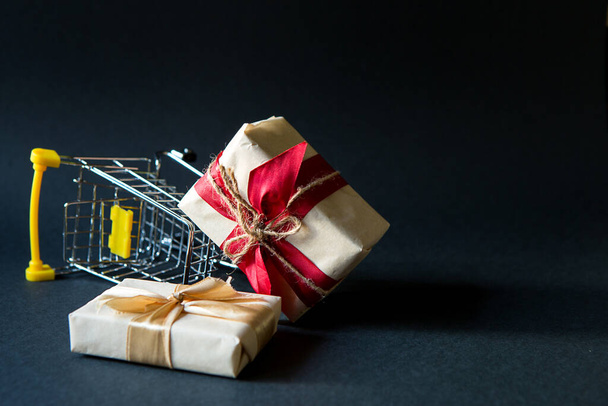 Box with a gift in Christmas packaging in a shopping cart on a black background. Black Friday, buying gifts for the new year. Space for text - 写真・画像