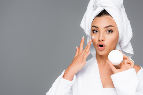 surprised woman with towel on head and cosmetic cream on face holding container isolated on grey - Foto, Imagen