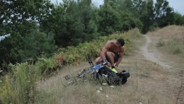 Um jovem homem musculoso de calções azuis agachamentos ao lado de sua bicicleta e mochila e amarra seus tênis antes de treinar na natureza, em seguida, levanta-se com um telefone na mão. Vista frontal. Close-up - Filmagem, Vídeo