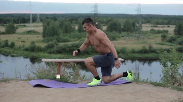 Un giovane uomo muscoloso con un busto nudo si allena vicino al fiume e fa oscillazioni delle gambe mentre sale su una panchina. Vista laterale. Primo piano. Camera si muove su e giù - Filmati, video