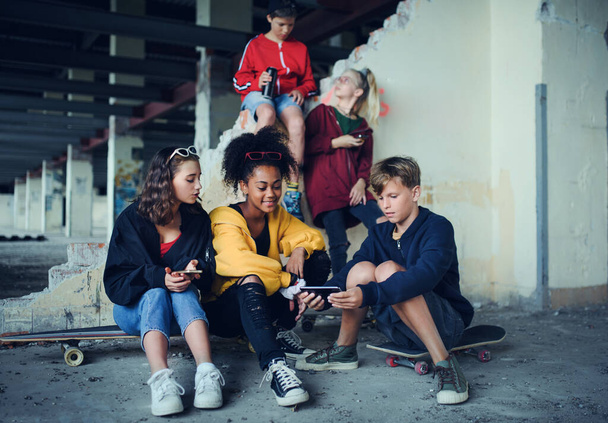 Group of teenagers gang sitting indoors in abandoned building, using smartphones. - Фото, изображение