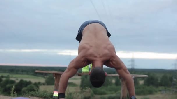 Ein junger muskulöser Mann mit nacktem Oberkörper legt seine Beine auf eine Bank und macht Liegestütze vor dem Hintergrund der Natur. Vorne. Nahaufnahme. Die Kamera bewegt sich auf und ab - Filmmaterial, Video