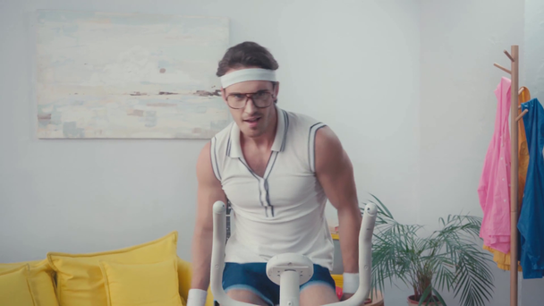 Sportsman in eyeglasses training on exercise bike in living room - Záběry, video