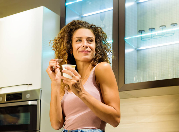 Boldog egészséges fiatal nő élvezi a kád friss joghurt a konyhában mosolyogva, ahogy néz félre egy alacsony szögből kilátás - Fotó, kép