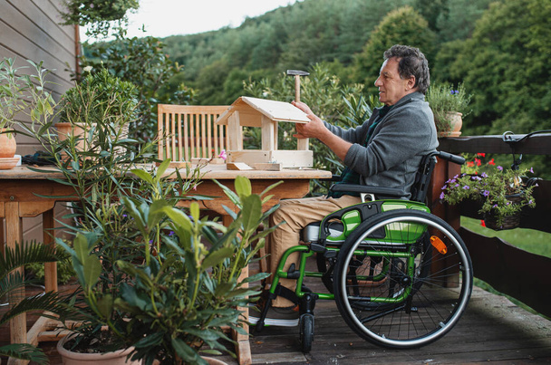 Senior man in wheelchair constructing birdhouse outdoors on terrace, diy project. - Foto, Imagem