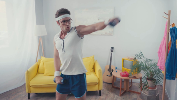 Спортсмен в очках поднимает гантели с протянутыми руками дома - Кадры, видео