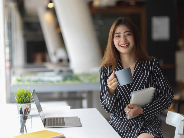 Portret van een lachend werkend vrouwelijk kantoor dat een koffiepauze neemt terwijl ze aan de werktafel zit  - Foto, afbeelding