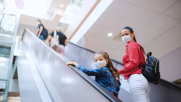Mother and daughter with face mask on escalator indoors in shopping center, coronavirus concept. - Photo, Image
