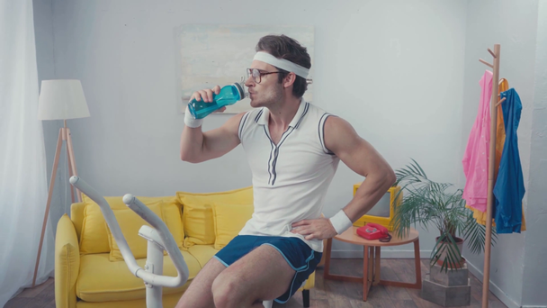 Müder Sportler trainiert auf Heimtrainer und trinkt Wasser im Wohnzimmer - Filmmaterial, Video