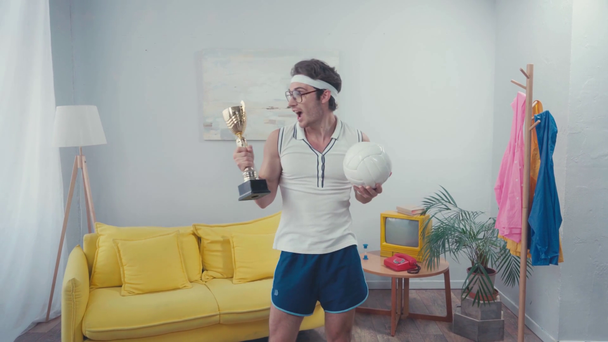Un sportif tenant une coupe trophée et une balle sportive, debout à la maison - Séquence, vidéo