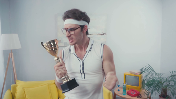 Sportsman with yeah gesture, celebrating win, while holding cup trophy at home - Footage, Video