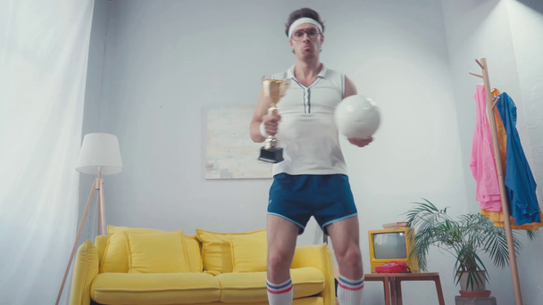 Enérgico deportista bailando con pelota deportiva y copa de trofeo en salón - Metraje, vídeo