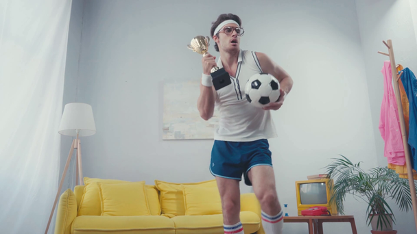 Энергичный футболист танцует со спортивным мячом и кубком в гостиной - Кадры, видео