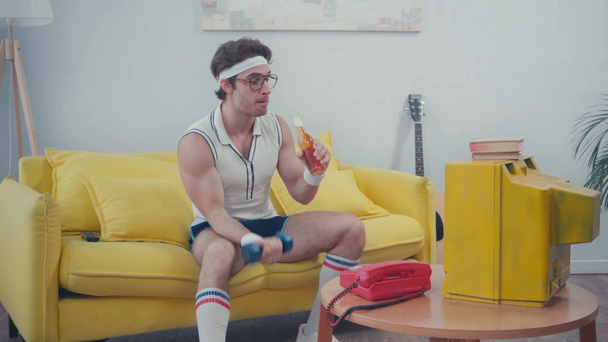 Urheilija juo juomaa ja nostaa käsipainoja istuessaan sohvalla - Materiaali, video