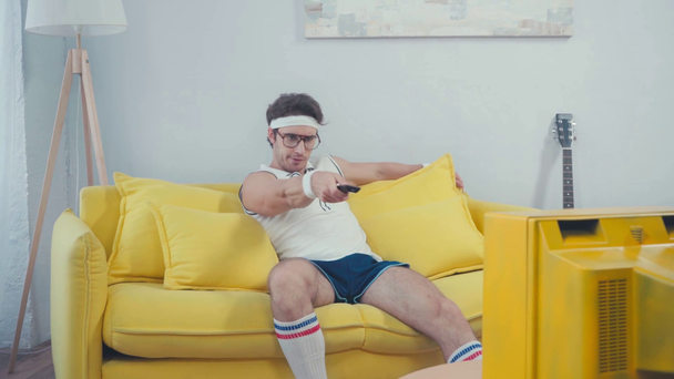 Lenivý muž ve sportovním oblečení spínací kanály, zatímco sedí na pohovce v obývacím pokoji - Záběry, video