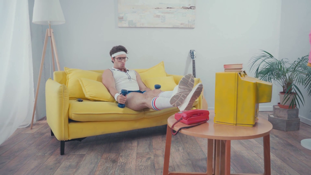Deportista con las piernas cruzadas en la mesa de café, levantando peso en la sala de estar - Metraje, vídeo