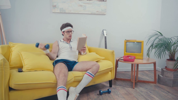 Urheilija nostaa käsipainoja, kun luet kirjaa sohvalla olohuoneessa - Materiaali, video