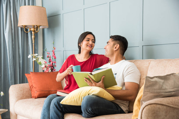 Young Indian asian couple reading books while sitting on sofa at home, modern living space - Foto, Bild