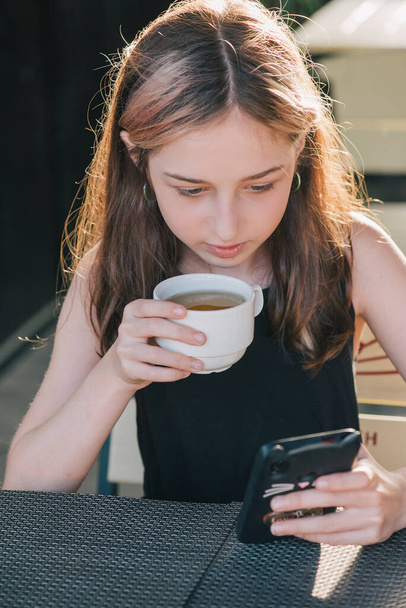 Nastolatka używająca telefonu komórkowego, przy filiżance herbaty shalllow DOF, kolorowy obraz. Nastolatka pije herbatę i patrzy na smartfona. - Zdjęcie, obraz