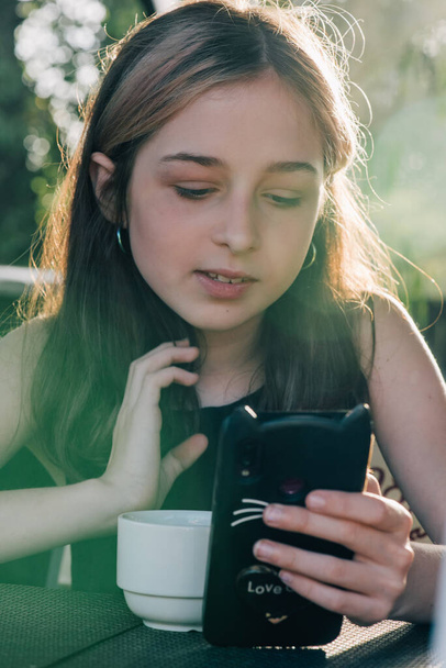 Teenage girl using her cell phone, while having a cup of tea shalllow DOF, color toned image. Teenage girl drinks tea and looks into a smartphone. - Photo, Image