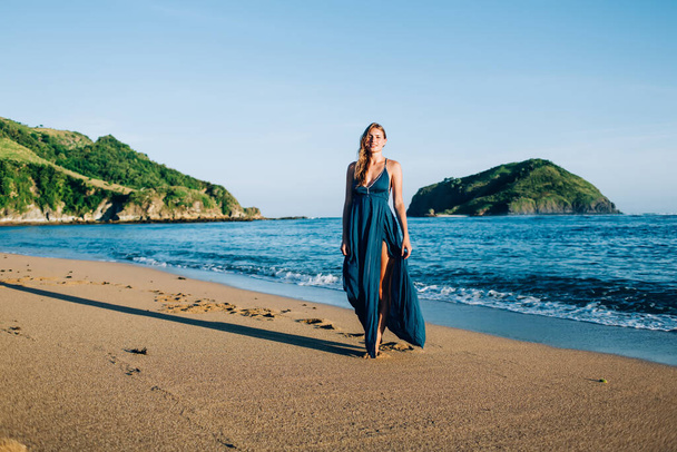 Beautiful relaxed blonde woman in long sundress strolling along idyllic sandy beach by bright blue waterfront and green hilly isles - Фото, изображение