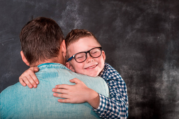 Little boy resting on her father's shoulder. Little son hugging his dad near wal. Happy father and child boy on hands. School kid boy in glasses. Father's day - Photo, Image