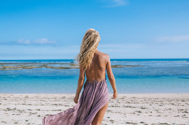 Back view of young blond female in lilac dress with open back looking at ocean while standing alone on tropical coastline in summertime - Foto, Bild