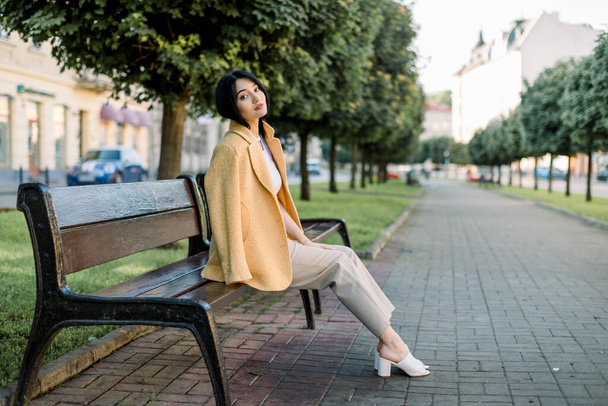Pretty young Asian business lady, wearing stylish smart casual yellow blazer and beige pants, sitting on a bench at city alley between green trees and enjoying start of sunny day - Foto, Imagen