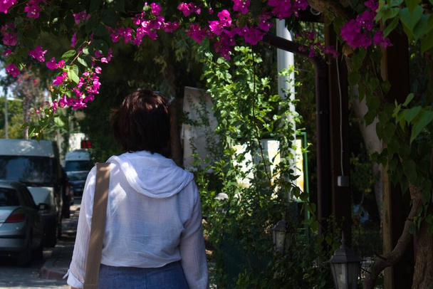 Frau in weißer heller Jacke auf einer schattigen Straße mit hellen Blumen an einem Sommertag - Foto, Bild