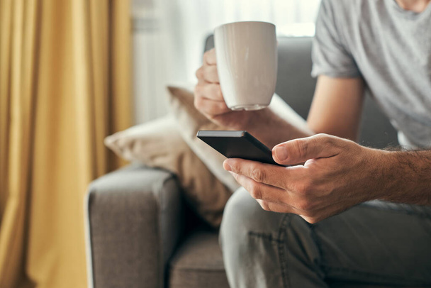 Man drinking coffee and using mobile phone at home, adult caucasian male enjoying morning routine at living room sofa - Photo, image
