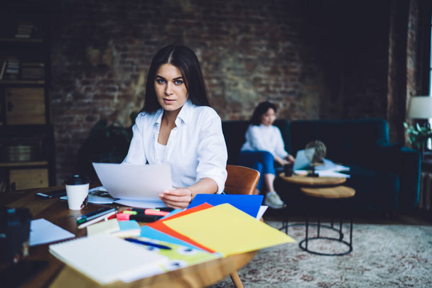 Concentrated female manager in casual outfit sitting at wooden table and looking at camera while working with documents in office - Фото, зображення