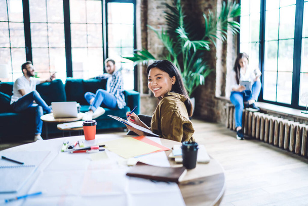 Happy Asian female in casual wear sitting at wooden table and taking notes in notebook while working with colleagues in loft office - Photo, Image