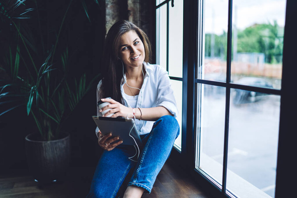 Young smiling female in casual outfit using tablet with earphones holding paper cup while relaxing on windowsill in modern workspace - Fotoğraf, Görsel