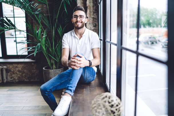 Full body of cheerful man employee sitting on windowsill and looking at camera while having break during work in modern workplace - Foto, Imagem