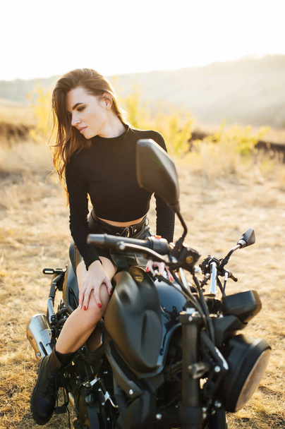 Beautiful girl in black clothes sits on motorcycle and poses for camera. Evening sunset - Valokuva, kuva
