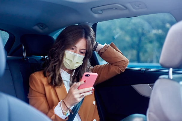 Белая женщина в маске для лица с помощью мобильного телефона в машине - Деловая женщина отправляет сообщение - Фото, изображение