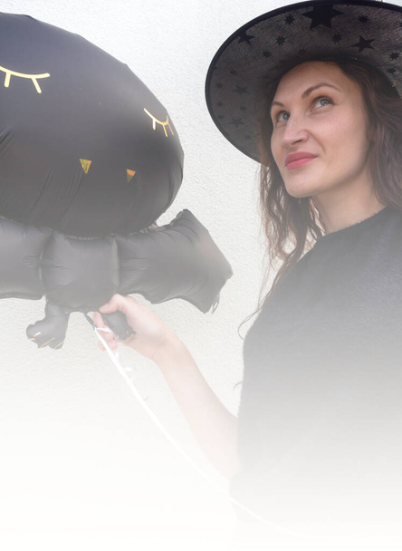 mujeres guapas vestidas con vestidos negros, sombrero de bruja y labios rojos sostienen bola de helio de gato de halloween negro, prepararse para la fiesta, tonificado con gradiente borroso blanco para su texto - Foto, Imagen
