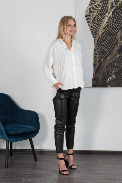 Stylish beautiful young blond woman in a tight black pants and white shirt near a white wall in the room. Attractive girl model posing. Fitness lady - Φωτογραφία, εικόνα