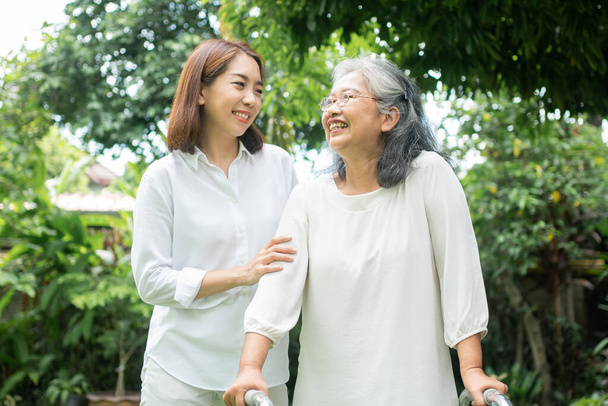 Een oude Aziatische vrouw gebruikt een looprek en loopt in de achtertuin met haar dochter. Concept van gelukkig pensioen Met zorg van een zorgverlener en spaargeld en senior ziektekostenverzekering - Foto, afbeelding