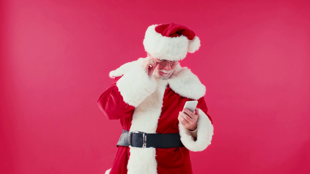 Ευτυχισμένος Άγιος Βασίλης κουβεντιάζοντας στο smartphone και αγγίζοντας γυαλιά που απομονώνονται στο κόκκινο - Πλάνα, βίντεο