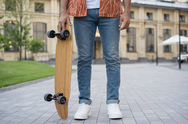 Стильний молодий чоловік у повсякденному одязі, біле взуття тримає скейтборд, стоїть у парку
 - Фото, зображення
