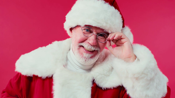 gioioso Babbo Natale toccando occhiali e sorridendo alla fotocamera isolata sul rosso - Filmati, video