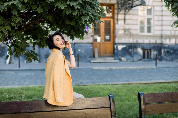 Laughing elegant Asian woman in yellow jacket, chilling and having fun during her morning city walk. Outdoor photo of lovely brunette business lady sitting on bench. - 写真・画像
