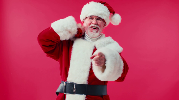 happy santa claus carrying sack and pointing with finger isolated on red - Footage, Video