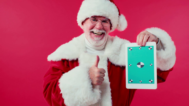 glimlachende kerstman wijst naar digitale tablet geïsoleerd op rood - Video