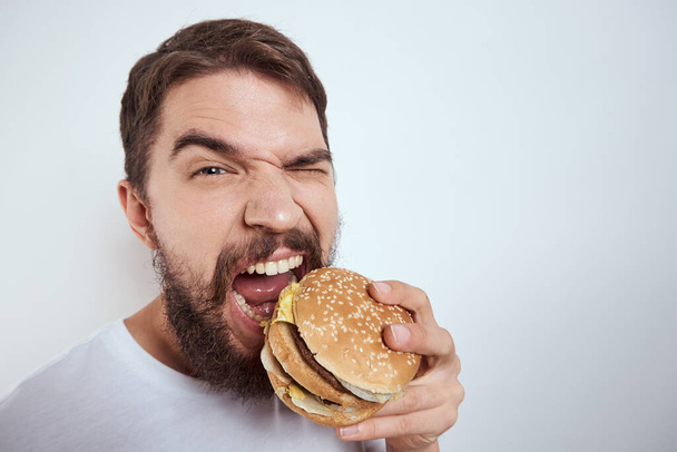 A man eating a hamburger on a light background in a white T-shirt cropped view close-up hunger fast food - Foto, imagen