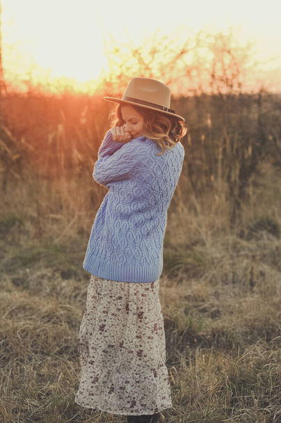Portrét mladé ženy v béžovém klobouku, modrém svetru a v květinových šatech relaxuje a užívá si slunce v teplém parku při západu slunce. Venkovní, štěstí - Fotografie, Obrázek