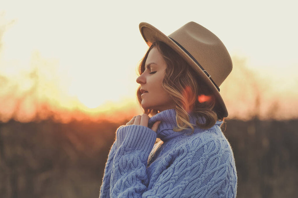 Портрет молодої жінки в бежевому капелюсі і синьому светрі розслаблює і насолоджується сонцем в теплому парку на заході сонця. Захід сонця - Фото, зображення