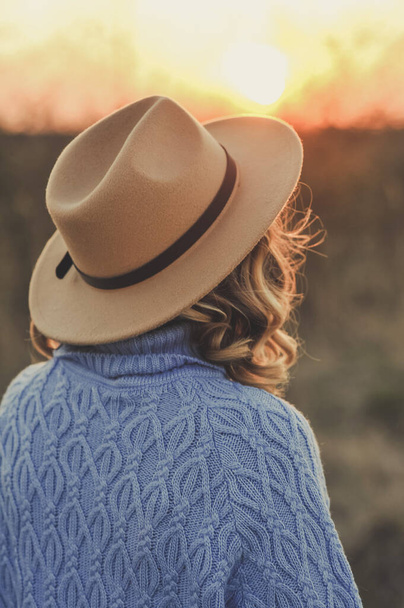 Vista posteriore di giovane donna in cappello beige e maglione blu guardando in lontananza al tramonto. All'aperto - Foto, immagini