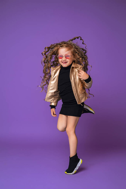 happy stylish girl 6 years old in fashionable clothes and sunglasses jumps in the air in full growth, isolated on a purple background. Dynamic emotional image. Flying in the air. Copy space. - Foto, Imagem